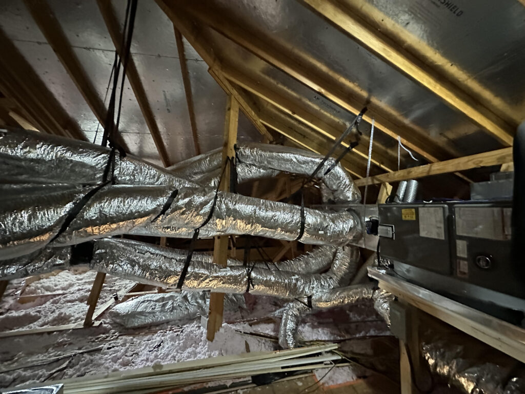 Installed ductwork in an attic, connected to an HVAC unit, with insulated ducts secured along the ceiling framework.