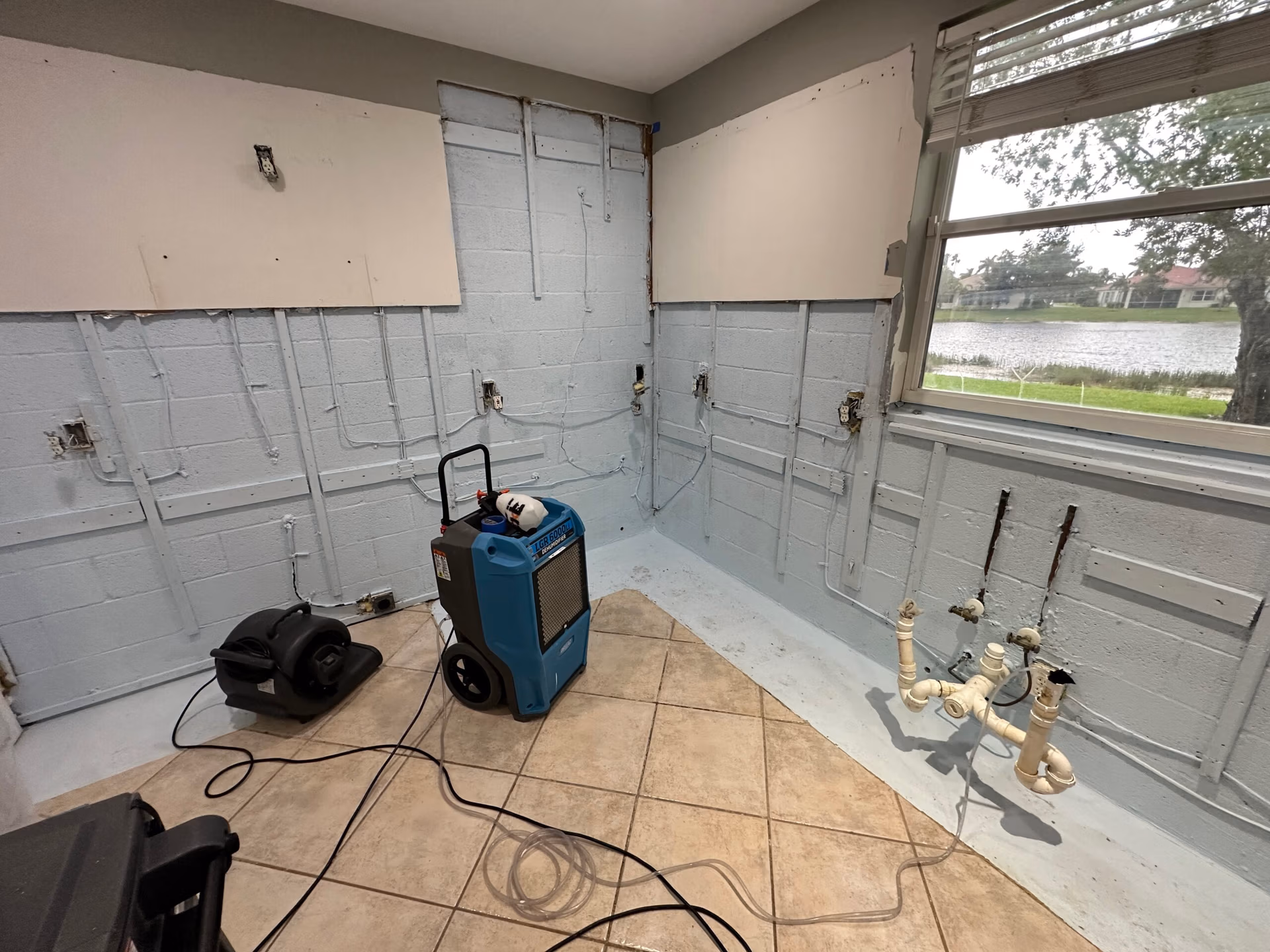 A partially demolished interior wall with exposed concrete blocks, electrical wiring, and plumbing fixtures, with dehumidifiers and air movers set up on the tile floor for drying.