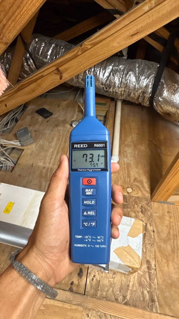 Technician holding a digital relative humidity meter in an attic to measure relative humidity levels near HVAC ductwork.