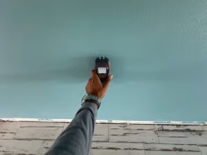 Hand holding a moisture meter against a painted wall, reading a high moisture level.
