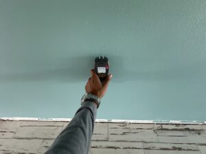 Hand holding a moisture meter against a painted wall, reading a high moisture level.