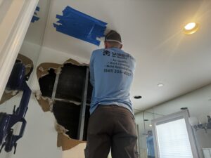 A restoration specialist in a blue shirt works on a damaged ceiling area with exposed insulation and framing, with plastic tape covering parts of the ceiling.