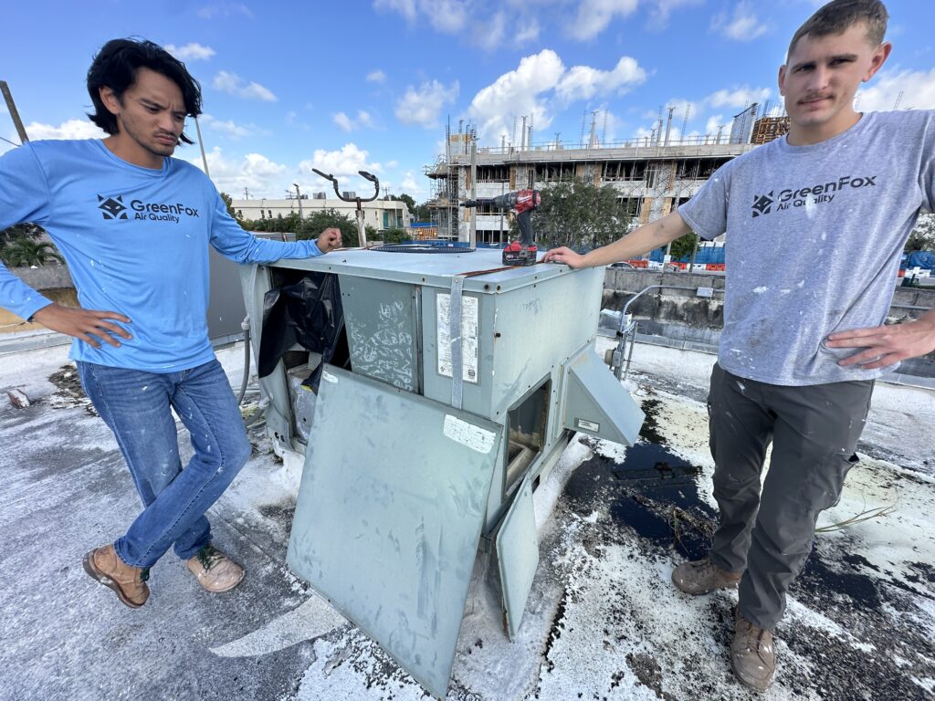 Two HVAC technicians from GreenFox Air Quality standing beside a rooftop air conditioning unit during maintenance work, with tools and an open access panel visible.