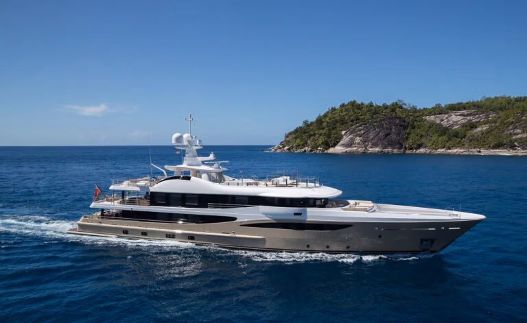 A luxury yacht cruising on clear blue waters with a small, forested island in the background. The yacht has a sleek, modern design with multiple decks, including antennas and navigation equipment on the top deck.