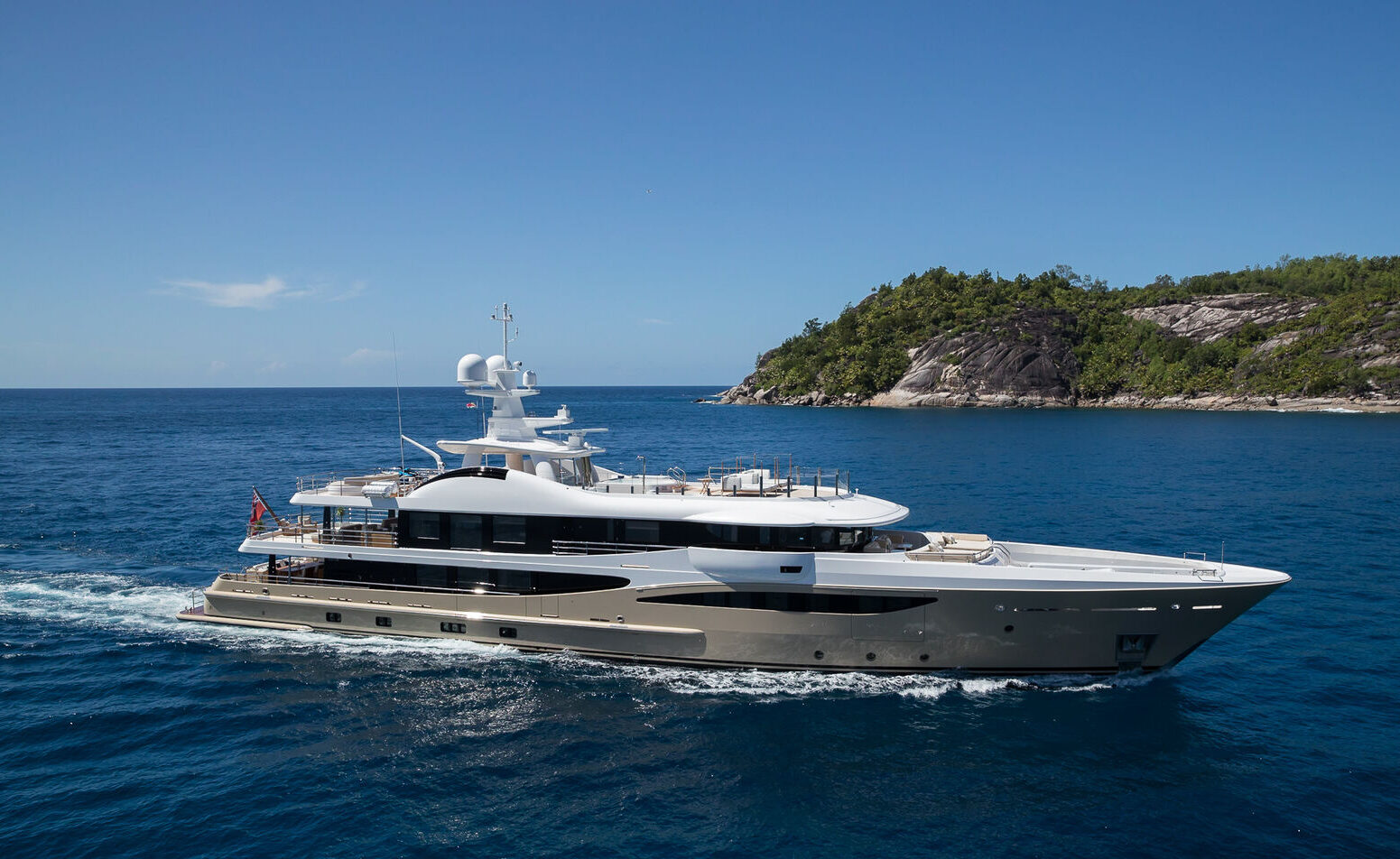 A luxury yacht cruising on clear blue waters with a small, forested island in the background. The yacht has a sleek, modern design with multiple decks, including antennas and navigation equipment on the top deck.