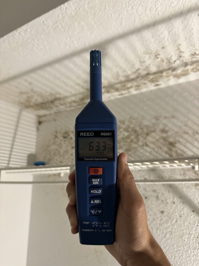 Hand holding a digital thermo-hygrometer showing 63.3% humidity near a ceiling with visible mold growth.