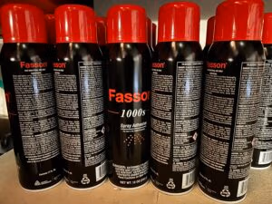 Black aerosol cans of Fasson 1000s spray adhesive designed for HVAC insulation and duct liner applications, arranged on a warehouse shelf.