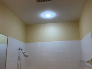 Striped wallpaper installed above white tile in a bathroom shower area with visible ceiling vent and recessed light.